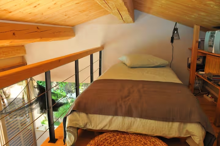 Lit simple situé en mezzanine sous un toit rampant en bois massif avec petite étagère de rangement.