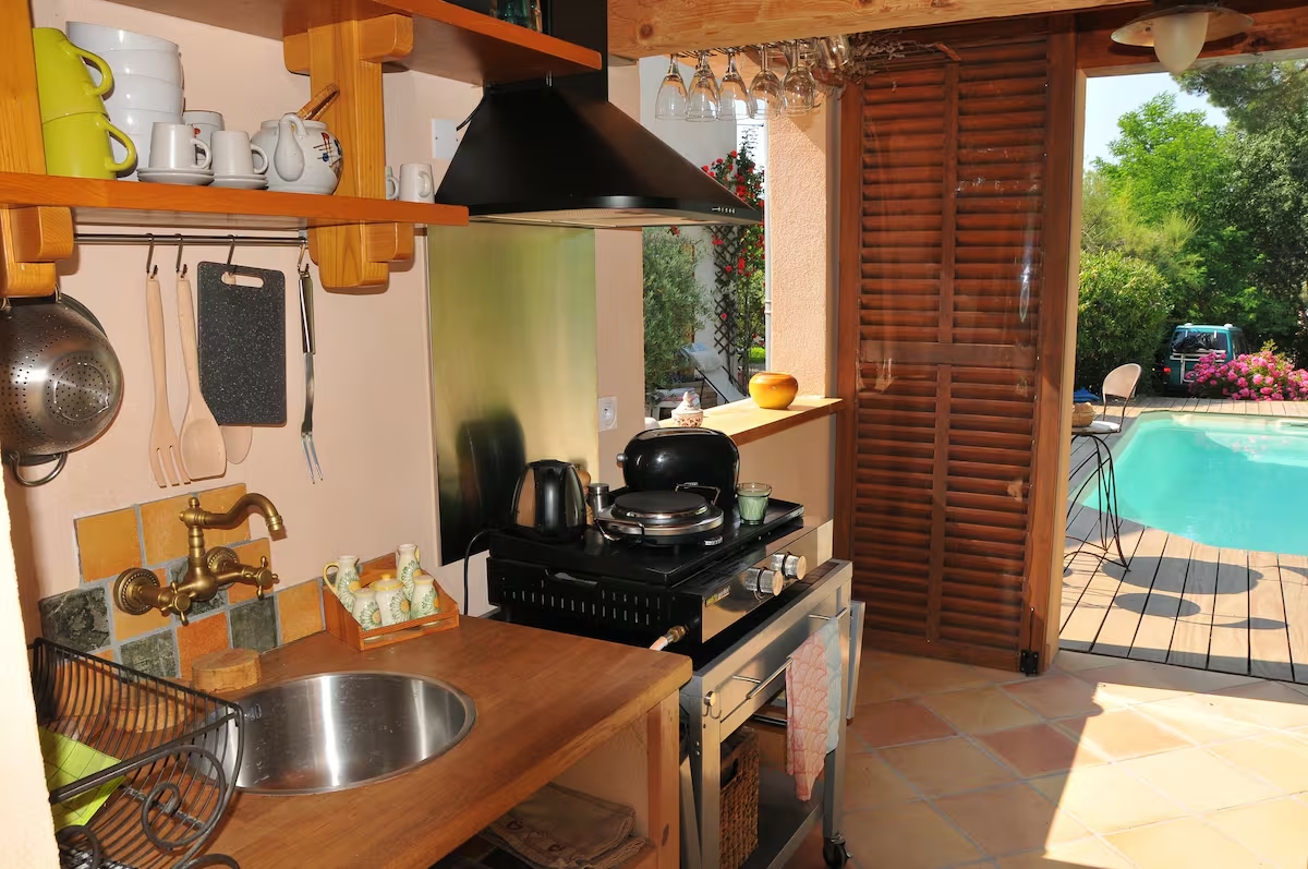 Cuisine d'été ouverte avec évier en inox, plaque de cuisson, étagères en bois garnies de vaisselle colorée et vue directe sur la piscine.