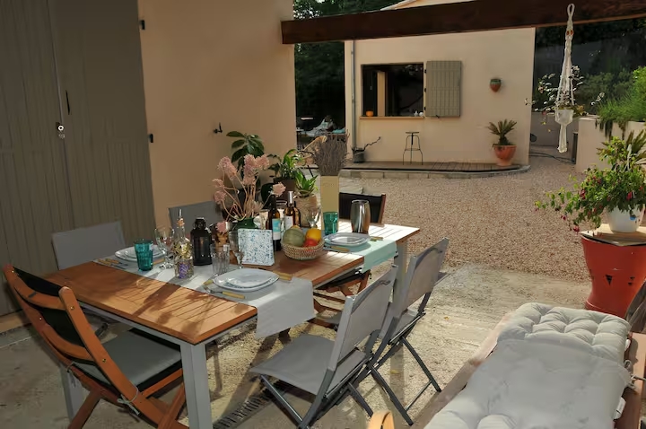 Table à manger extérieure couverte, dressée pour plusieurs convives dans une cour en gravier avec plantes vertes.