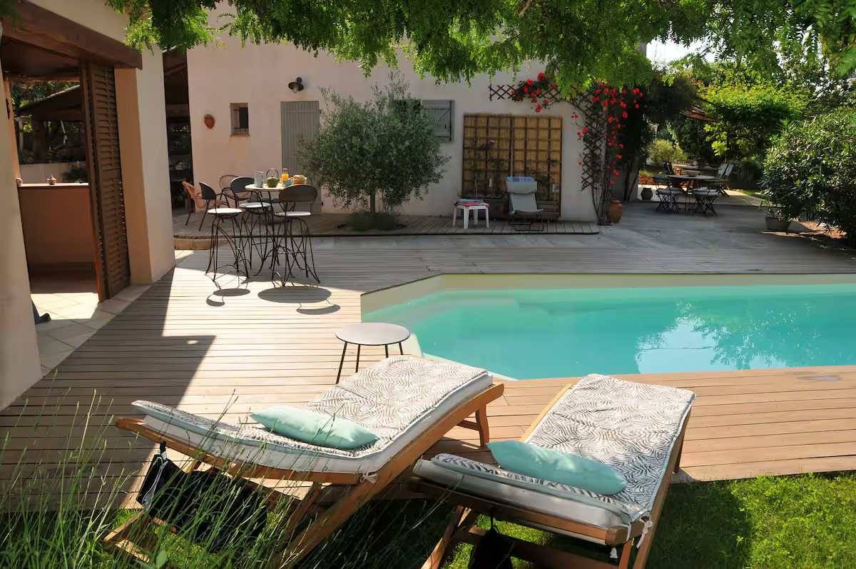 Espace détente avec chaises longues au bord de la piscine, terrasse ombragée et façade de maison provençale avec fleurs grimpantes rouges.