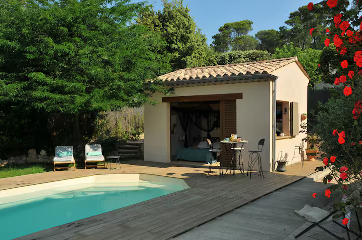 Façade d'un pool house en pierre avec toit en tuiles provençales, table de bistrot sur la terrasse et piscine en premier plan sous un soleil radieux.